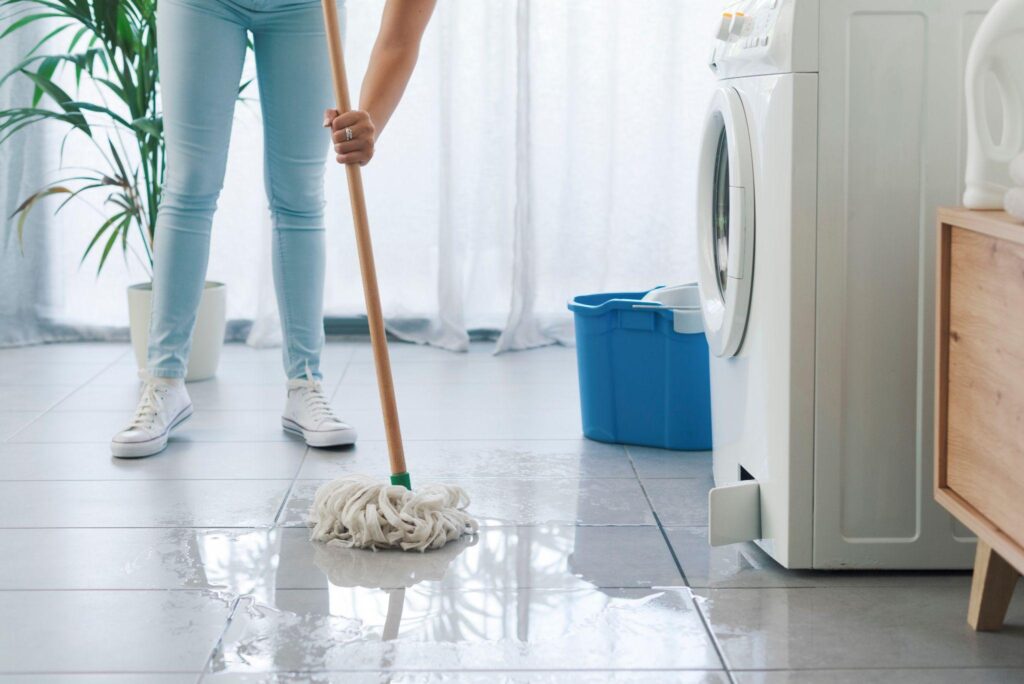 A person mopping the floor in front of a washing machine.